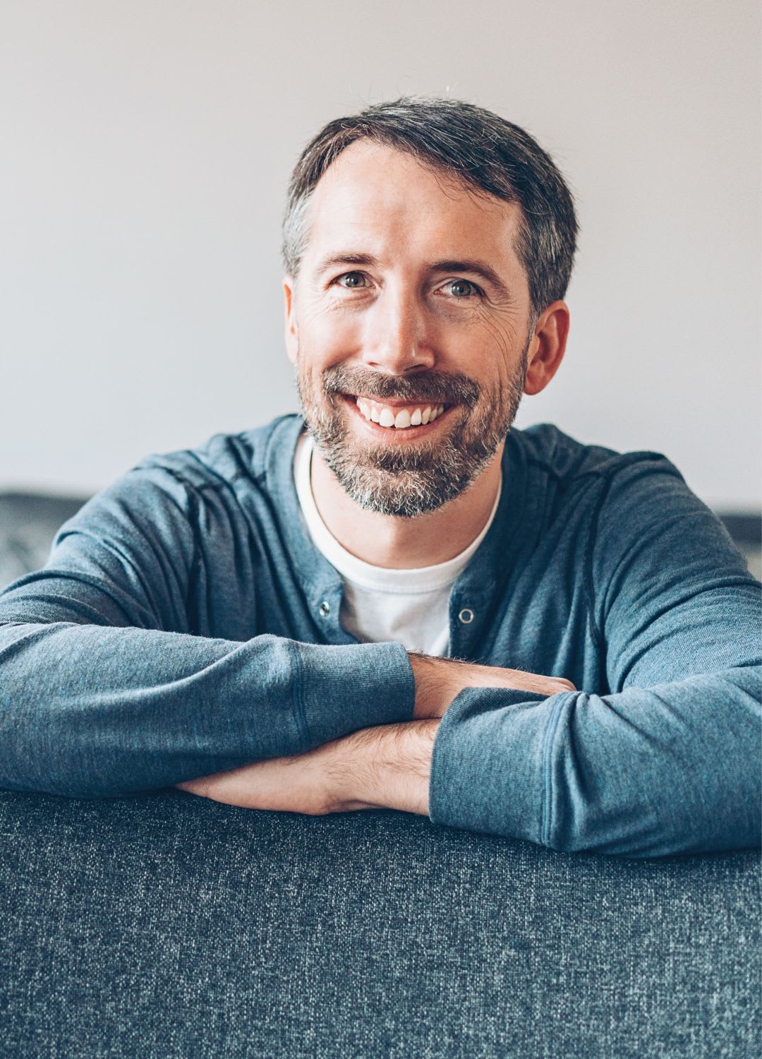 A man with short brown hair and a beard, wearing a blue sweater, smiles while sitting on a gray sofa with his arms crossed on the backrest after his ivig infusion.